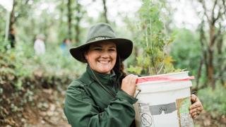 Smiling woman outdoors in raingear and hat holding a white bucket with tree sapling