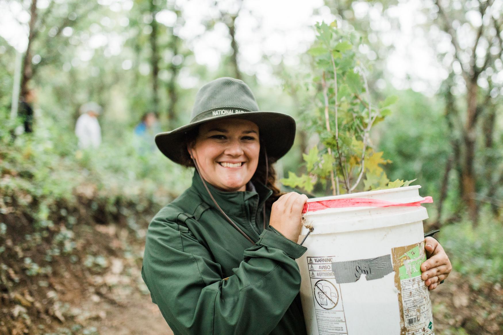 Smiling woman outdoors in raingear and hat holding a white bucket with tree sapling 